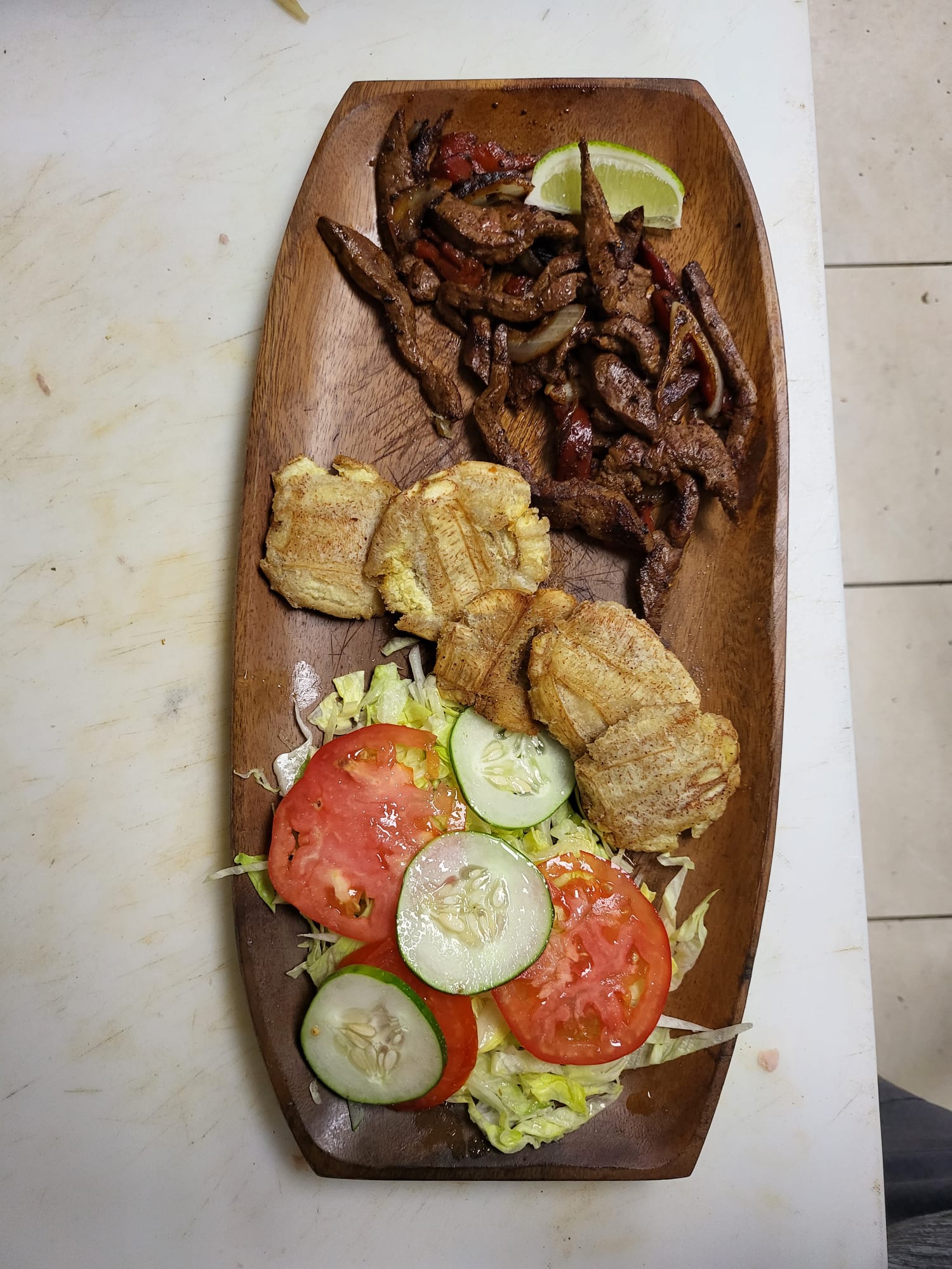 Overhead shot of a rustic wooden platter featuring sautéed beef strips with onions and peppers, fried plantains, and a fresh salad.